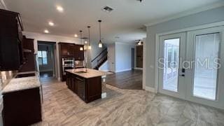 kitchen featuring dark brown cabinetry, ornamental molding, an island with sink, decorative light fixtures, and french doors