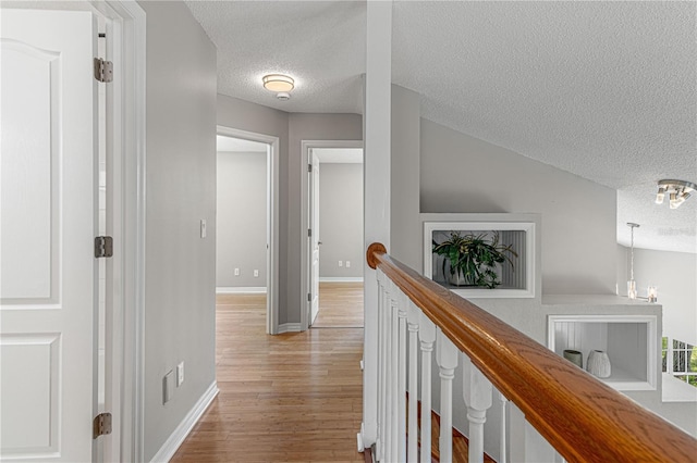 corridor featuring light wood-type flooring, baseboards, a textured ceiling, and an upstairs landing