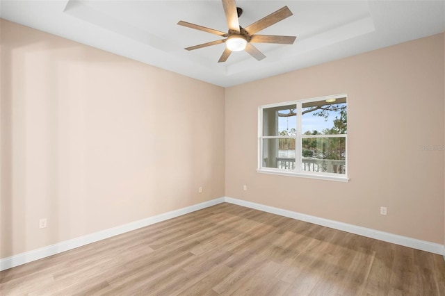 empty room with ceiling fan, light wood finished floors, a raised ceiling, and baseboards