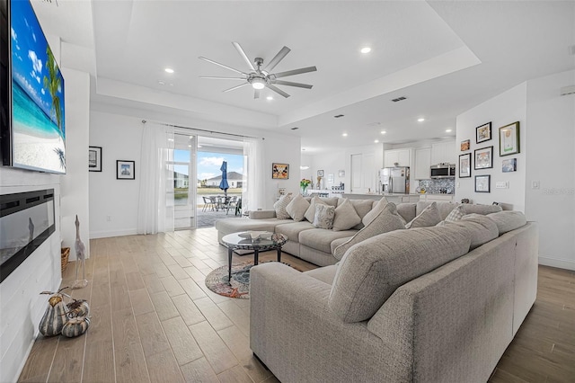 living room featuring a tray ceiling, light hardwood / wood-style flooring, a large fireplace, and ceiling fan