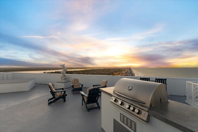 patio terrace at dusk with exterior kitchen, a water view, and a grill