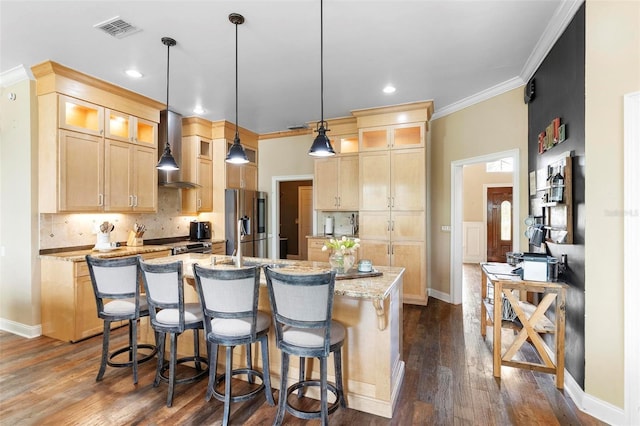 kitchen featuring appliances with stainless steel finishes, light brown cabinetry, light stone counters, wall chimney range hood, and a kitchen island with sink