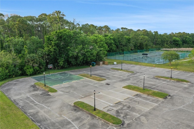 view of tennis court featuring community basketball court, a wooded view, and fence