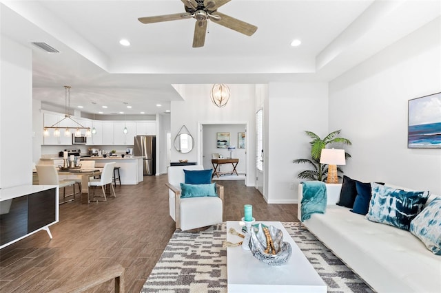 living room with dark wood-type flooring, ceiling fan with notable chandelier, and a raised ceiling