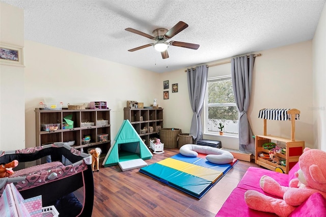 recreation room with a ceiling fan, a baseboard radiator, a textured ceiling, and wood finished floors