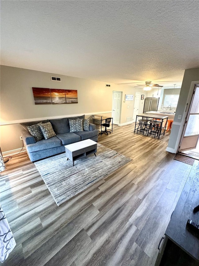 living room with a textured ceiling, ceiling fan, wood finished floors, visible vents, and baseboards