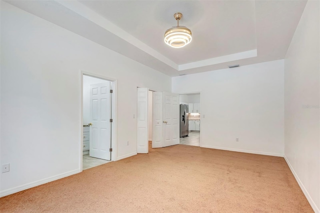 unfurnished bedroom with light carpet, stainless steel fridge, a raised ceiling, and ensuite bathroom