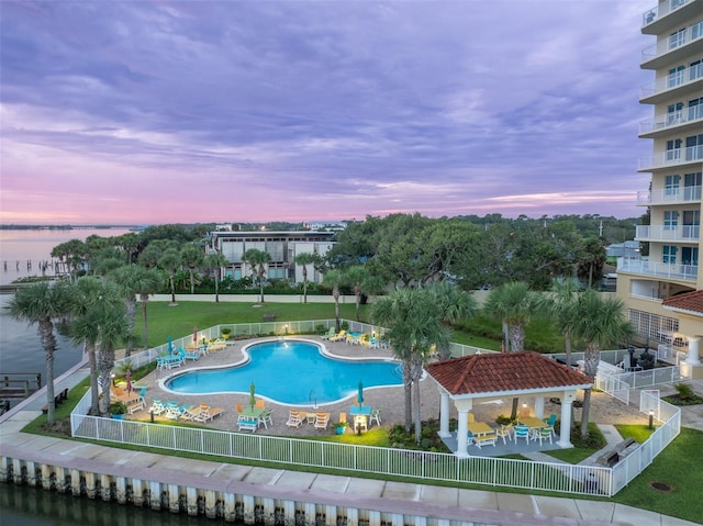 pool at dusk with a water view, a gazebo, a patio area, fence, and a community pool