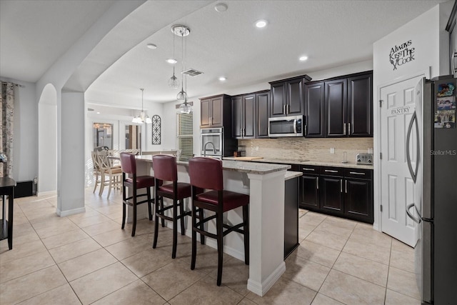 kitchen featuring arched walkways, light tile patterned floors, a kitchen island with sink, appliances with stainless steel finishes, and backsplash