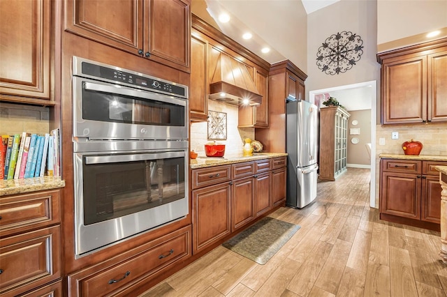 kitchen featuring custom range hood, appliances with stainless steel finishes, brown cabinets, light wood-style floors, and backsplash