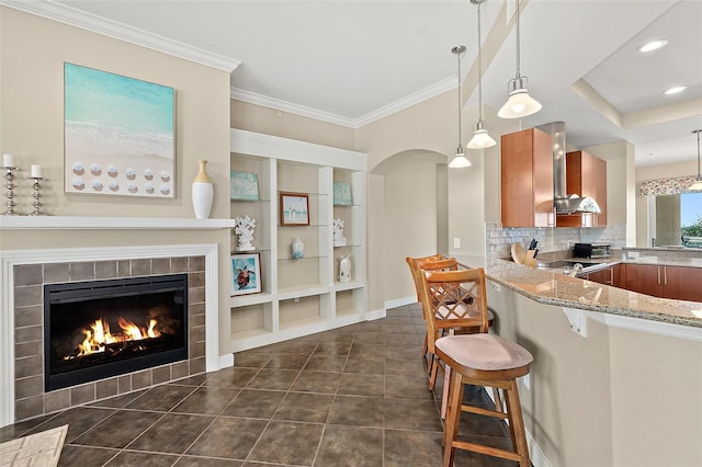 kitchen featuring light stone counters, a fireplace, wall chimney exhaust hood, brown cabinetry, and a kitchen bar