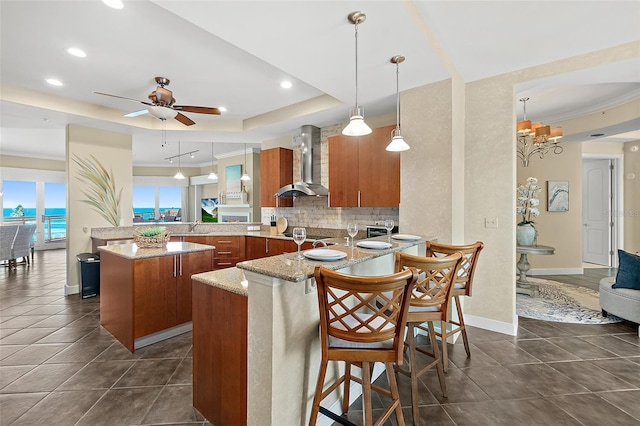 kitchen featuring a peninsula, wall chimney range hood, backsplash, a tray ceiling, and brown cabinetry