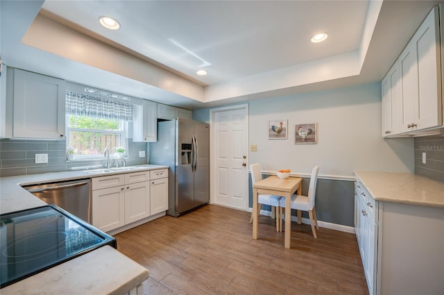 kitchen with stainless steel appliances, a raised ceiling, decorative backsplash, a sink, and light wood-type flooring