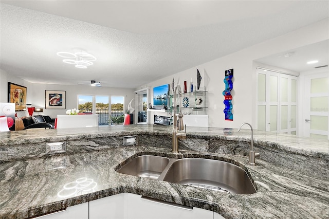 kitchen with a textured ceiling, ceiling fan, a sink, open floor plan, and dark stone countertops