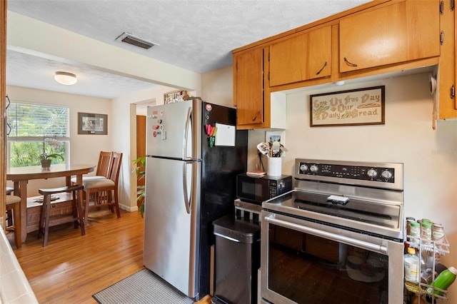 kitchen with a textured ceiling, visible vents, light wood-style floors, appliances with stainless steel finishes, and brown cabinetry