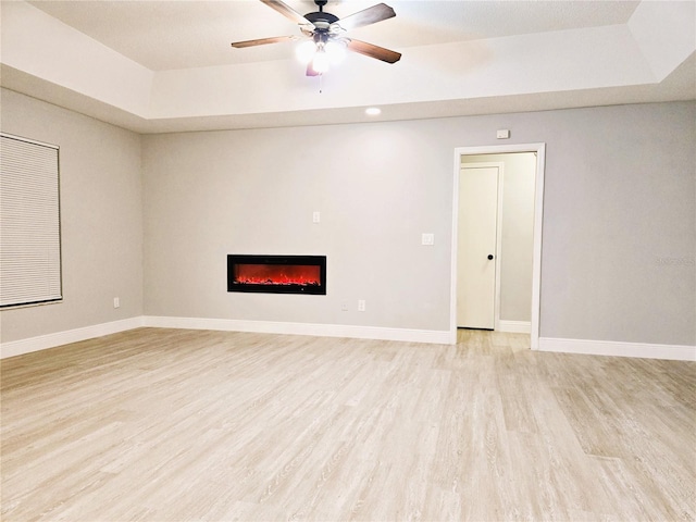 unfurnished living room with ceiling fan, baseboards, light wood-style floors, a raised ceiling, and a glass covered fireplace