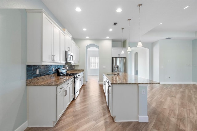 kitchen featuring arched walkways, light wood-style flooring, a kitchen island with sink, stainless steel appliances, and backsplash