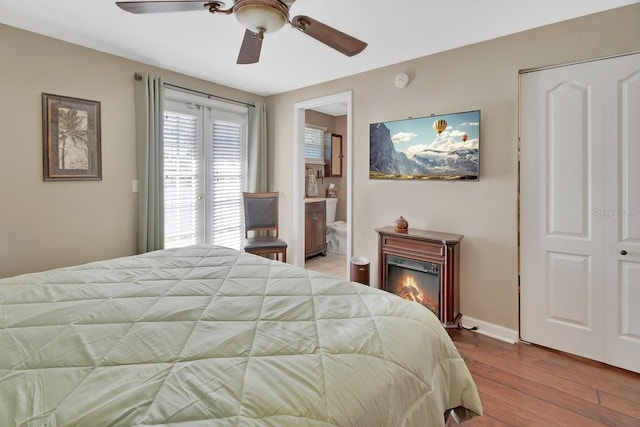 bedroom featuring baseboards, a ceiling fan, a glass covered fireplace, ensuite bath, and wood finished floors