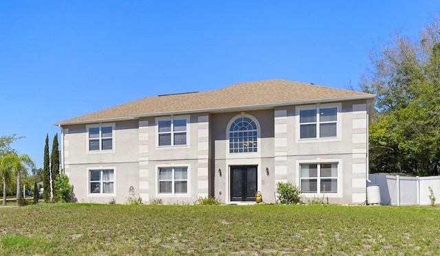 colonial-style house with stucco siding, fence, french doors, roof with shingles, and a front yard