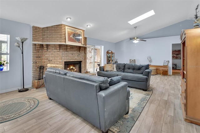 living room featuring ceiling fan, lofted ceiling with skylight, a brick fireplace, and light wood-style floors