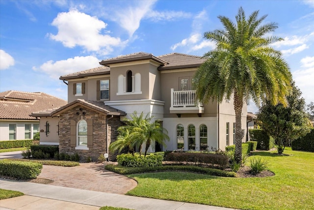 mediterranean / spanish-style house featuring a front yard, a balcony, stucco siding, stone siding, and a tile roof