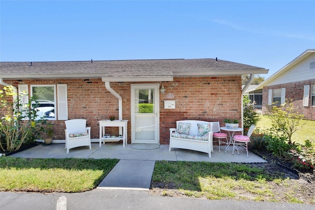 doorway to property featuring brick siding, a shingled roof, an outdoor hangout area, a yard, and a patio