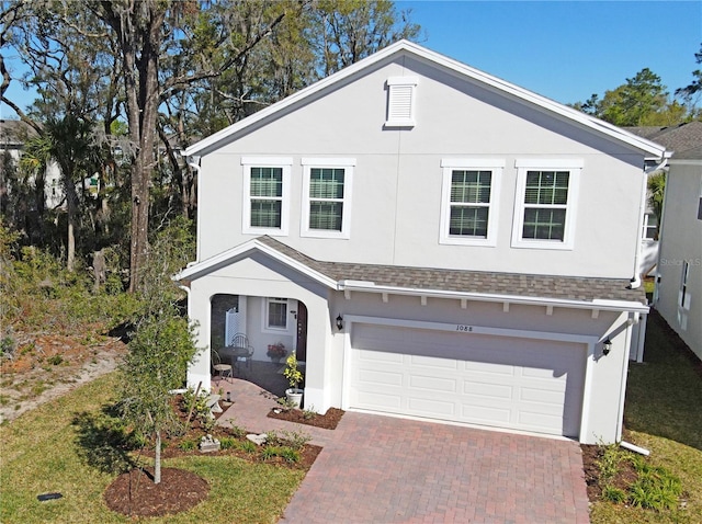 view of front of home with decorative driveway, roof with shingles, an attached garage, and stucco siding