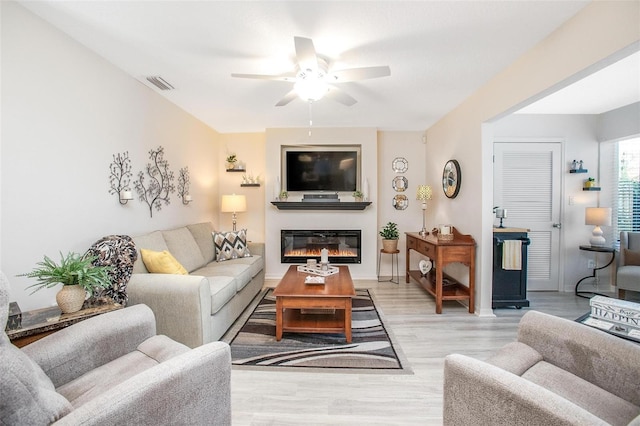 living room featuring baseboards, visible vents, ceiling fan, a glass covered fireplace, and light wood-type flooring