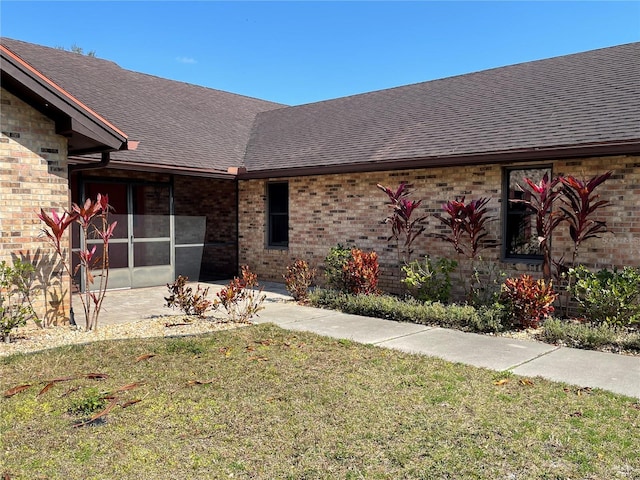 doorway to property featuring a patio, a yard, brick siding, and roof with shingles