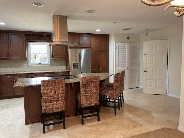 kitchen with stainless steel refrigerator with ice dispenser, light stone counters, a kitchen breakfast bar, island range hood, and decorative backsplash