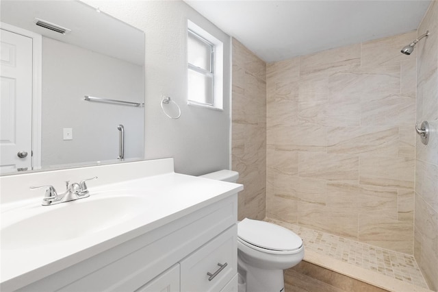 bathroom featuring visible vents, toilet, wood finished floors, tiled shower, and vanity