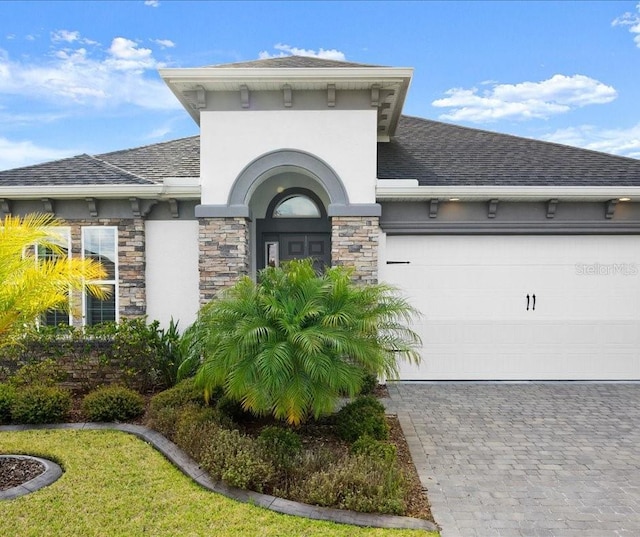exterior space with decorative driveway, stone siding, an attached garage, stucco siding, and roof with shingles