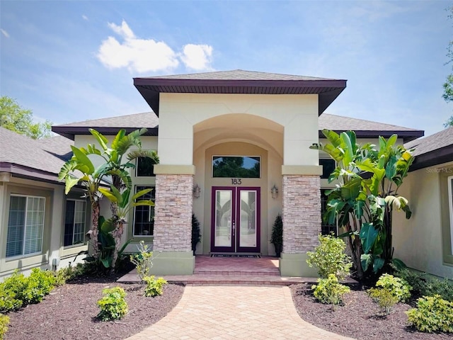 view of exterior entry with a shingled roof, stucco siding, french doors, and stone siding