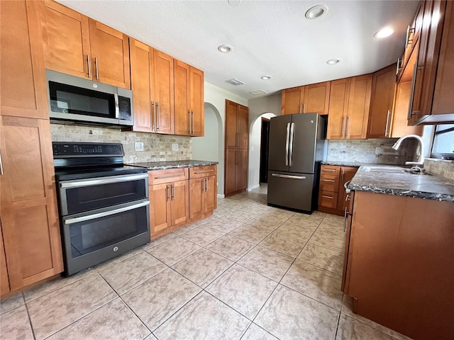 kitchen featuring arched walkways, decorative backsplash, brown cabinetry, stainless steel appliances, and a sink