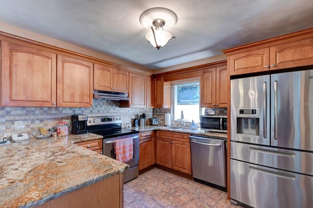 kitchen with light stone counters, stainless steel appliances, under cabinet range hood, brown cabinetry, and backsplash