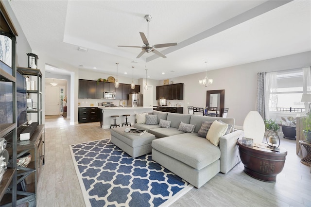 living room with a raised ceiling, ceiling fan with notable chandelier, recessed lighting, and light wood-style flooring