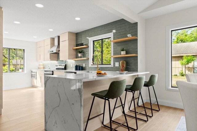 kitchen with open shelves, a sink, a peninsula, light wood-style floors, and wall chimney exhaust hood