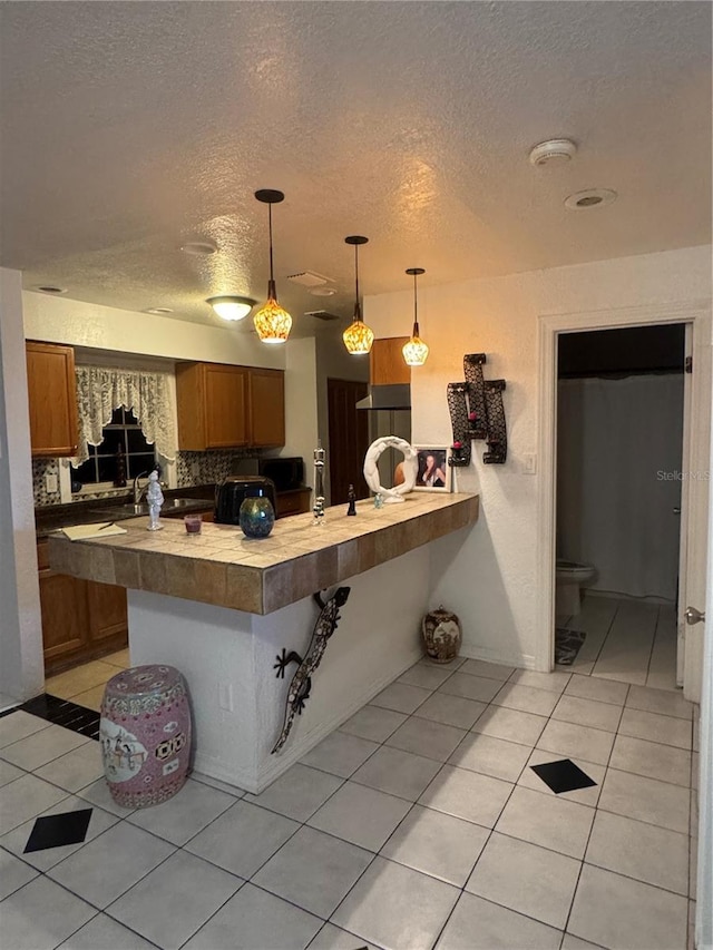 kitchen featuring brown cabinets, a textured ceiling, light tile patterned floors, a peninsula, and hanging light fixtures