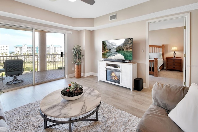 living area featuring a ceiling fan, a glass covered fireplace, light wood-type flooring, and visible vents