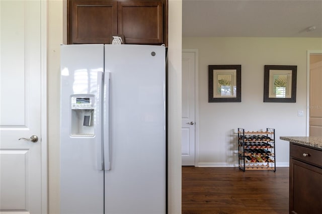 kitchen featuring white refrigerator with ice dispenser, dark wood-style floors, dark brown cabinetry, light stone counters, and baseboards