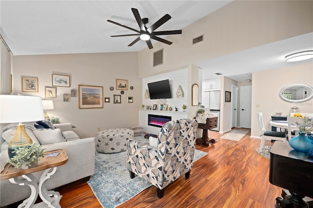 living room with a ceiling fan, wood finished floors, a glass covered fireplace, and high vaulted ceiling