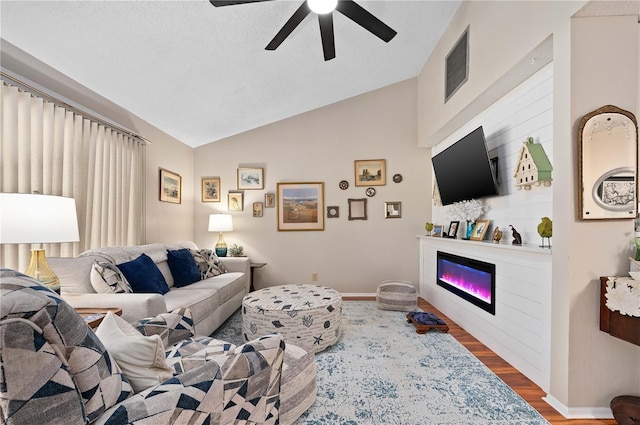 living area featuring wood finished floors, a glass covered fireplace, a ceiling fan, lofted ceiling, and baseboards