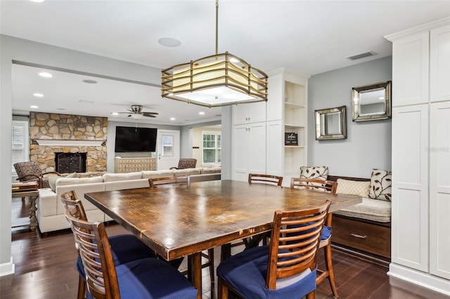 dining area with a stone fireplace, ceiling fan, and dark wood-type flooring