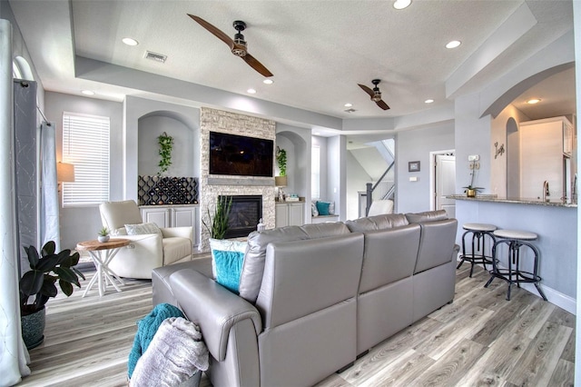 living room with light wood-type flooring, a textured ceiling, a raised ceiling, ceiling fan, and a stone fireplace