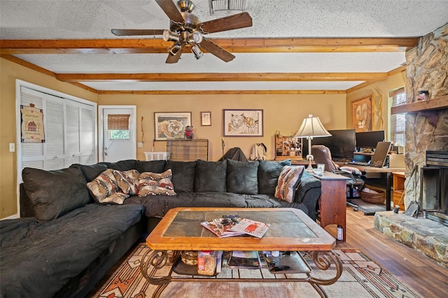 living room with beam ceiling, hardwood / wood-style flooring, a stone fireplace, ceiling fan, and a textured ceiling