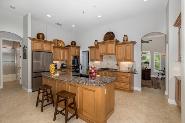 kitchen featuring backsplash, a kitchen island with sink, dark stone counters, ceiling fan, and stainless steel refrigerator