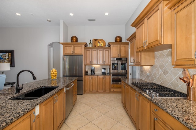 kitchen with appliances with stainless steel finishes, backsplash, dark stone counters, sink, and light tile patterned floors