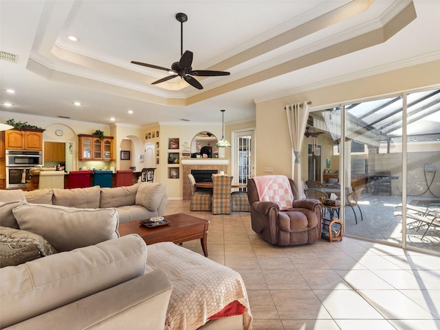 living room featuring a raised ceiling, ceiling fan, ornamental molding, a fireplace, and light tile patterned floors