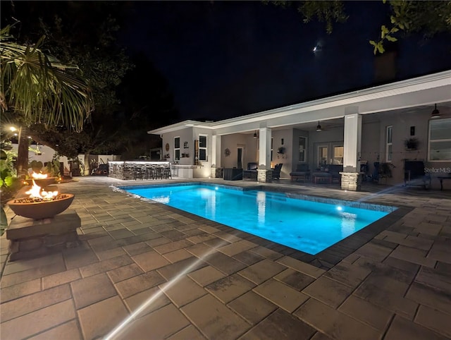 pool at night featuring a fire pit, a patio, exterior bar, and a ceiling fan