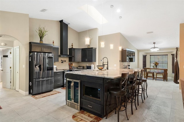 kitchen featuring wall chimney exhaust hood, lofted ceiling, light stone counters, appliances with stainless steel finishes, and a kitchen breakfast bar